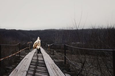 Man in raincoat standing on footbridge against sky