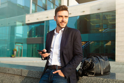 Smiling young man using mobile phone while standing against office building 