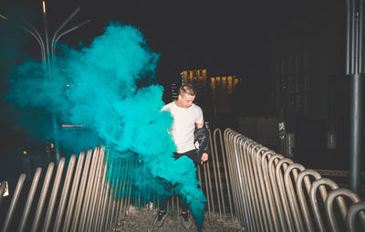 Full length of young man standing by distress flare amidst metallic railings at night