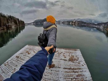 Cropped hand of man holding woman on pier over lake against cloudy sky