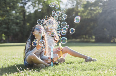 Two caucasian girls sisters blowing out a lot of soap bubbles while sitting in the park