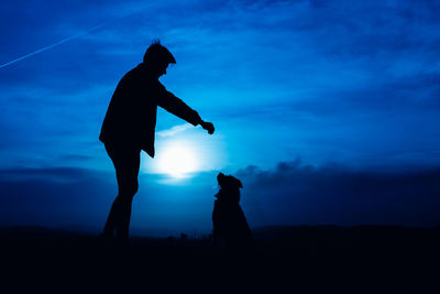 Silhouette of a young man feeding his dog with blue night light