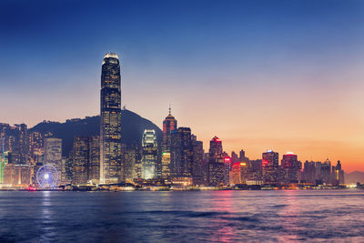 Victoria harbor against illuminated buildings in city at sunset