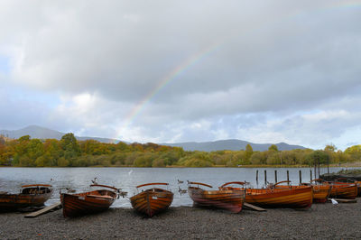 Boats moored on riverbank against rainbow in cloudy sky