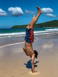 Shirtless man doing handstand at beach against sky