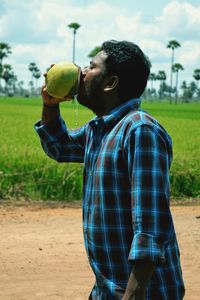 Side view of man drinking coconut water while standing on footpath by grassy field