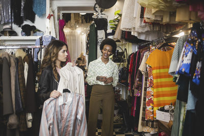 Young female customer trying on shirts while standing by owner in boutique