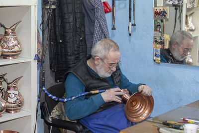 Senior man carving on brass container while sitting in workshop