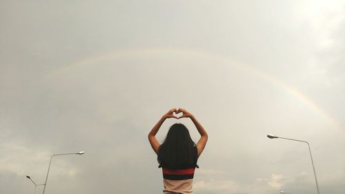 Low angle view of woman making heart shape while standing against rainbow in cloudy sky