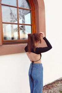 Back view of young woman standing against window raising up hands doing her hair