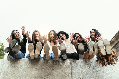 Portrait of happy friends gesturing while sitting on concrete wall against clear sky