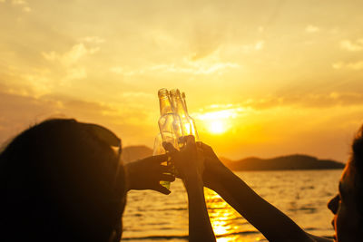 Friends toasting bottles while partying at beach against sky during sunset