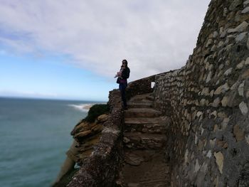 Woman standing on steps at mountain by sea against cloudy sky