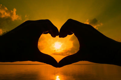 Cropped image of silhouette hand making heart shape against sky during sunset