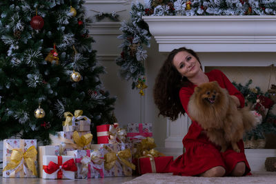 Portrait of smiling woman holding dog while sitting by christmas presents at home