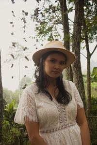 Portrait of young woman wearing hat standing against trees