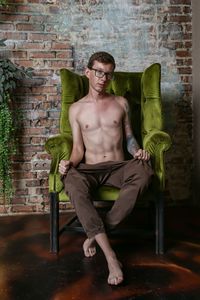 Portrait of shirtless man sitting against brick wall