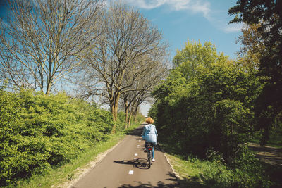 Rear view of woman riding bicycle on road against sky during sunny day