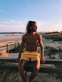 Shirtless man sitting on railing at beach against sky