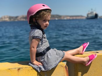 Cute girl wearing pink helmet sitting on retaining wall against sea during sunny day