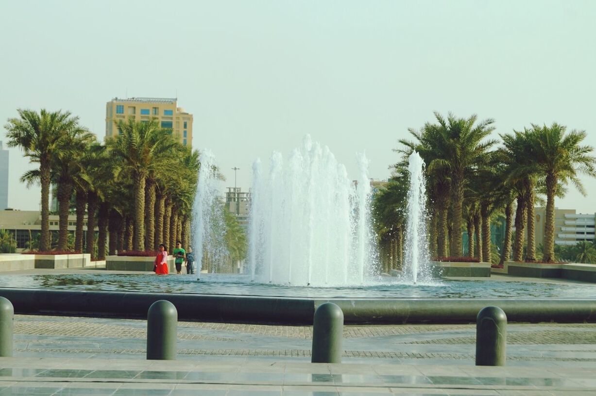 water, tree, clear sky, palm tree, built structure, architecture, building exterior, fountain, railing, incidental people, motion, city, day, sky, sea, outdoors, transportation, copy space, nature, sunlight