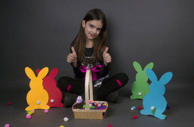 Portrait of smiling girl with eater bunny and pom pom in basket against gray background