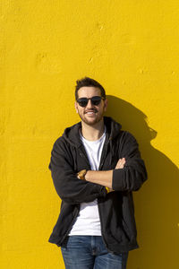 Young bearded man standing against a yellow wall with crossed arms