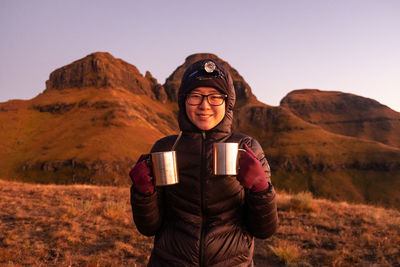 Lady in warm clothing smiling at camera with torch on her head and two steel cups at sunrise 