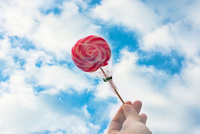 Low angle view of cropped hand holding lollipop against cloudy sky