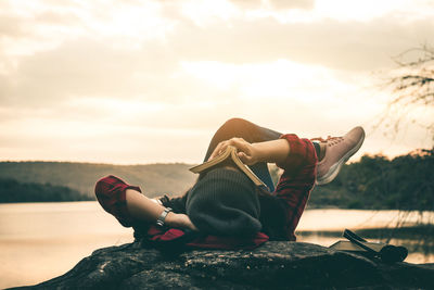 Woman resting on rock by lake against sky during sunset