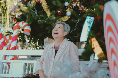 Cheerful senior woman sitting against christmas tree