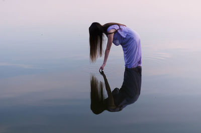 Side view of woman touching water in lake against sky during sunset