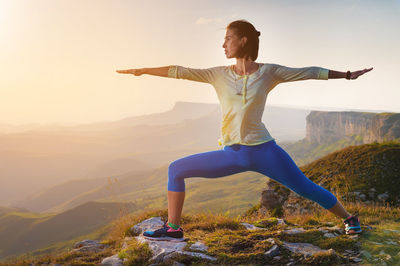 Yoga warrior pose of young caucasian woman practicing at sunset in the mountains in the grass. zen