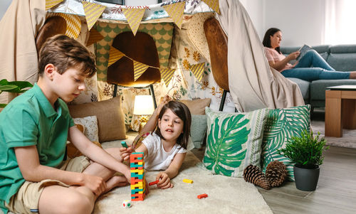 Little girl putting piece of stacking game while play with child and woman reading book on sofa