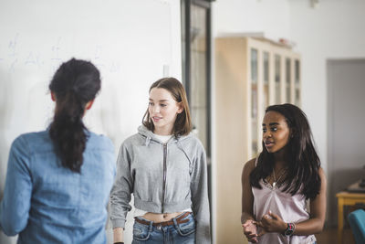 Rear view of teacher explaining female students standing by whiteboard in classroom at high school