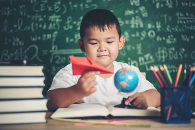 Cute boy studying at table against blackboard