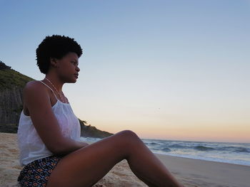 Young woman looking away while sitting on beach against sky during sunset