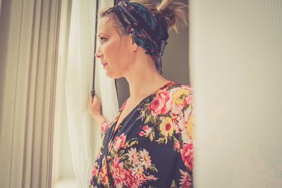 Young woman looking away while standing against window at home