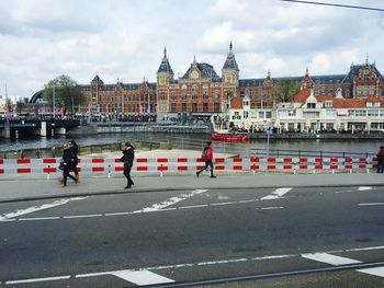 People walking on road by amsterdam centraal railway station against cloudy sky