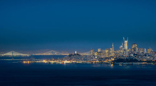 Illuminated city by sea against clear blue sky at night