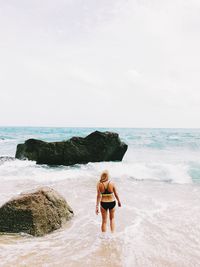 Rear view of woman standing by rock at beach against sky