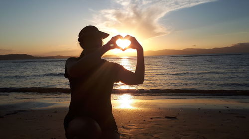 Silhouette woman making heart shape on beach against sky during sunset