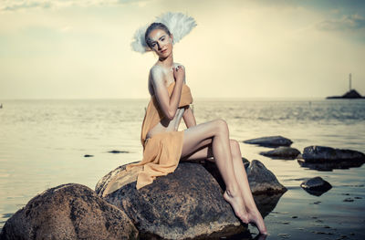 Portrait of female model wearing feathers while sitting on rocks in sea