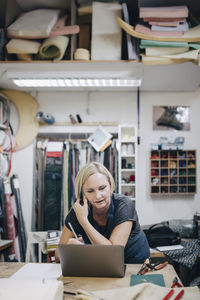 Mature female upholstery worker looking at laptop while talking on phone in workshop