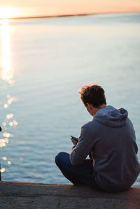 Man sitting on retaining wall against sea during sunset