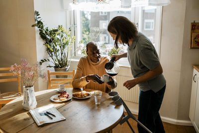 Side view of female nurse serving coffee to senior man having breakfast at home