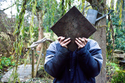 Man covering face with wood against trees