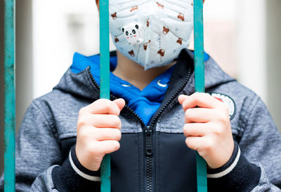 Close-up of child standing behind the gate and wearing face mask due to coronavirus pandemic.