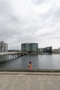 Rear view of woman sitting by river in city against sky