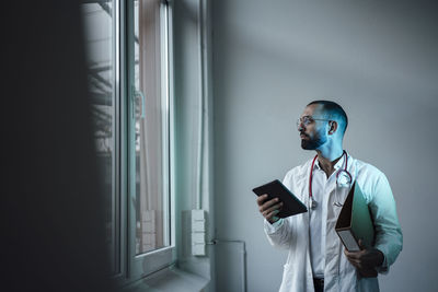 Thoughtful doctor holding tablet pc and file folder looking through window in front of wall at hospital